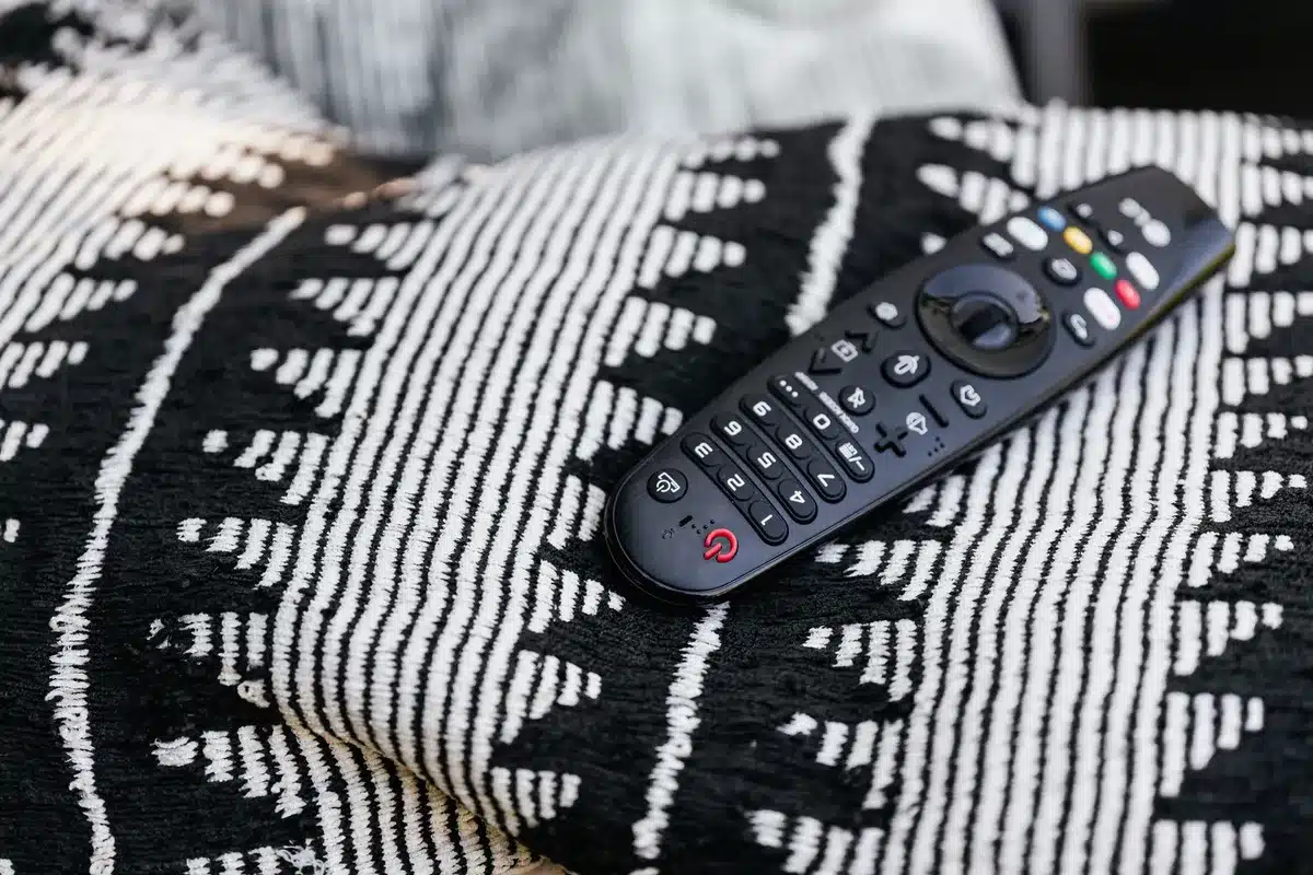 Close-up of a generic TV remote lying on a coffee table, anonymous hands reaching toward it, modern sofa in background; no branding visible